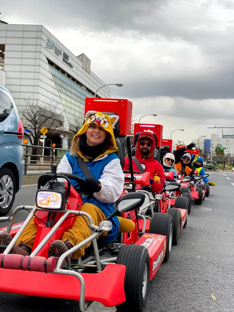 People riding red go-karts in a parade on a city street; front driver wears a yellow tiger hoodie and goofy smile under cloudy skies