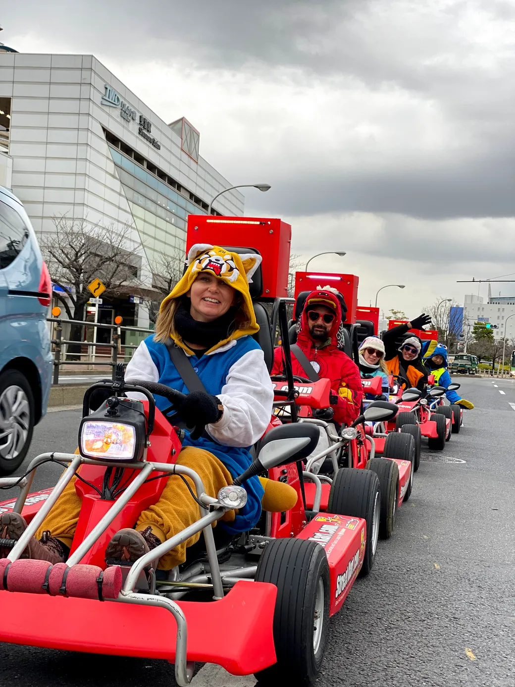People riding red go-karts in a parade on a city street; front driver wears a yellow tiger hoodie and goofy smile under cloudy skies