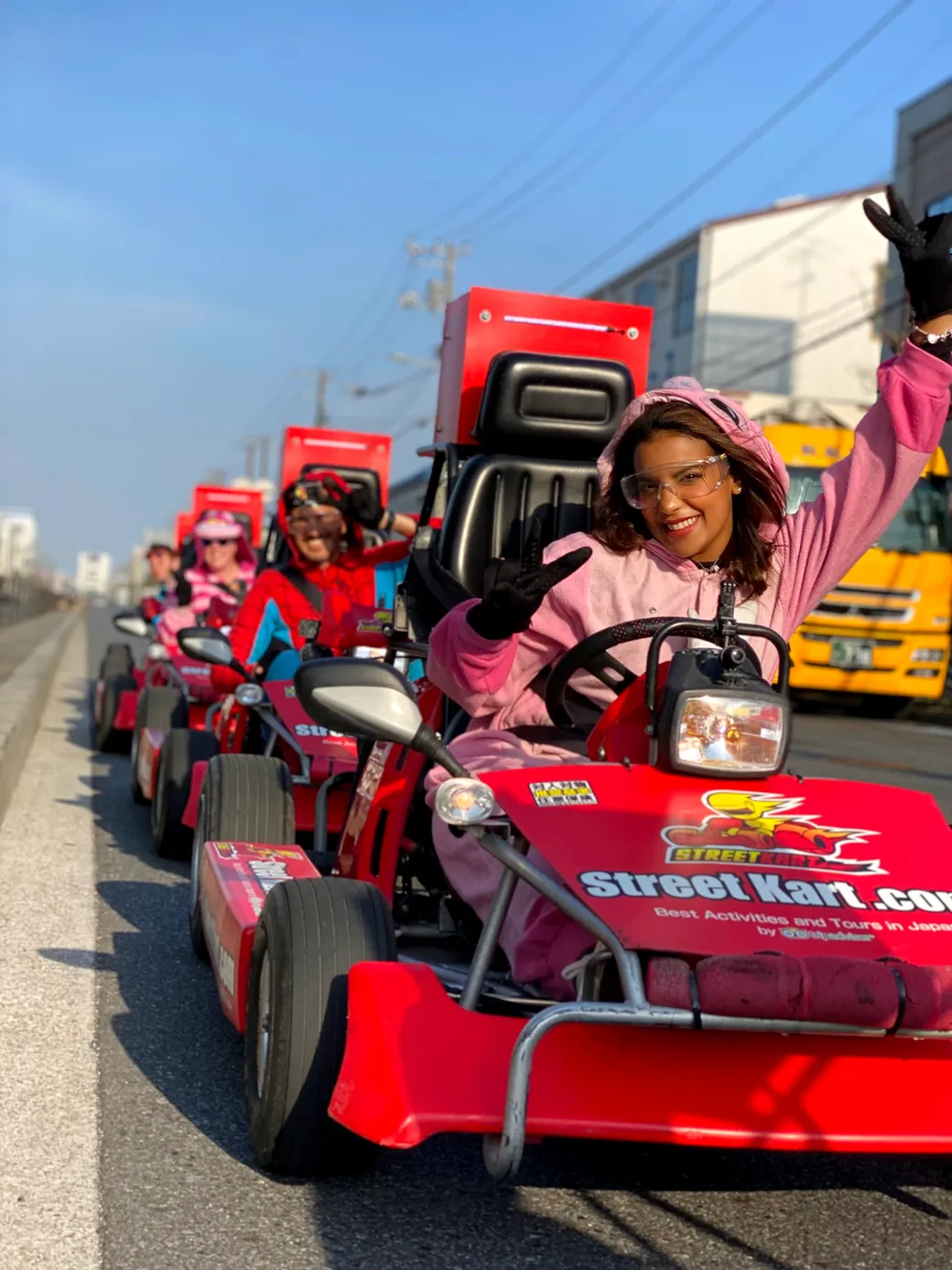 Smiling woman in a pink hoodie driving a red street-kart, waving from a line of go-karts on a city street with trucks in the background.