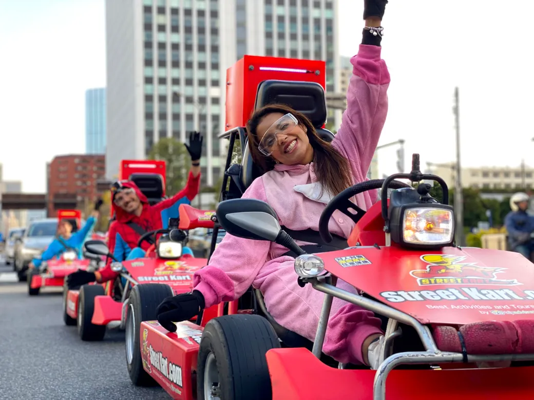 Group of people riding red go-karts in a city parade, smiling and waving to the crowd